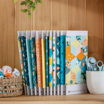 Twelve Missouri Star Bitty Bolts wrapped in teal, orange and yellow quilting fabric, standing upright on a wood shelf beside thread and rotary cutters